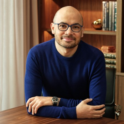 Demis Hassabis sits smiling with his arms crossed on a wooden desk, wearing a blue sweater and glasses.